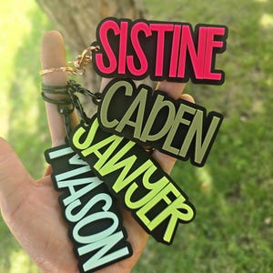 May include: Four rectangular keychains with black borders and bold lettering. The top keychain features the name SISTINE in hot pink, followed by CADEN in olive green, SAWYER in lime green, and MASON in light mint green, each attached to a metal ring.