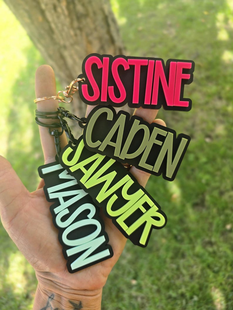 May include: Four rectangular keychains with black borders and bold lettering. The top keychain features the name SISTINE in hot pink, followed by CADEN in olive green, SAWYER in lime green, and MASON in light mint green, each attached to a metal ring.