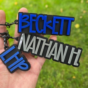 May include: Three rectangular keychains with black bases and raised lettering. The top keychain features the name BECKETT in blue, the middle displays NATHAN H. in silver, and the bottom shows the letters LHP in blue, all attached to black metal rings.