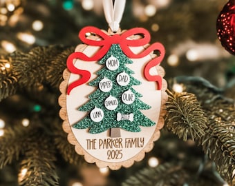 May include: An oval wooden Christmas ornament featuring a green glittery tree with white circular ornaments labelled Mom, Dad, Piper, Olive, Samuel, Liam, and a bone shape labelled Bella. A red bow sits atop the tree, with The Parker Family 2025 below.