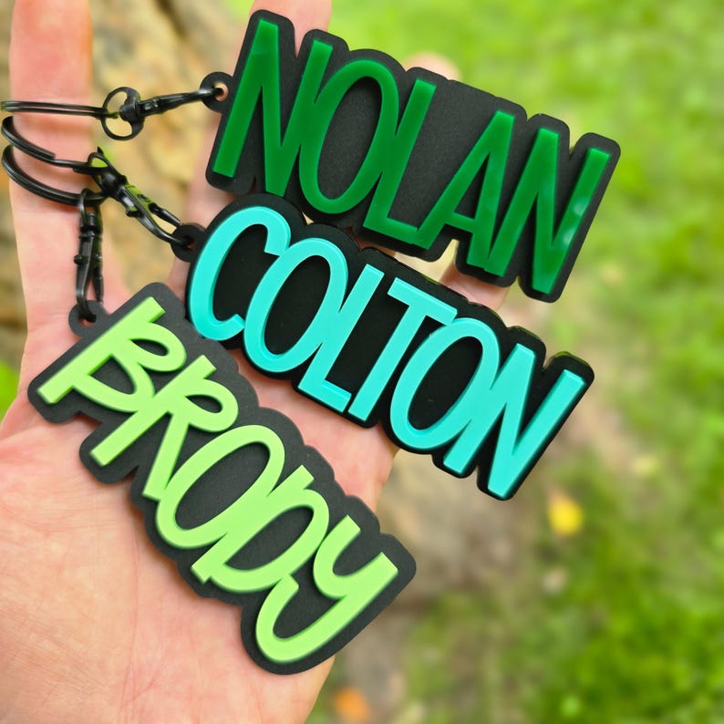 May include: Three custom keychains featuring the names NOLAN, COLTON, and BRODY in raised lettering. Each keychain has a black base with green, teal, and light green text respectively, attached to black metal rings with small black lobster clasps.