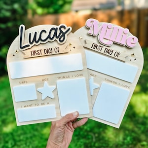 May include: Two wooden first day of school signs, one with "Lucas" in black and the other with "Millie" in pink. Each sign has space for date, age, and things I love. The signs are held by a person.