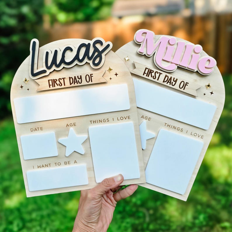 May include: Two wooden first day of school signs, one with "Lucas" in black and the other with "Millie" in pink. Each sign has space for date, age, and things I love. The signs are held by a person.