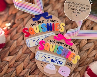 Könnte beinhalten: Zwei runde Holzornamente mit regenbogenfarbenen Streifenbändern. Jedes zeigt den Text 'the year of the SQUISHIES 2026', eine Grafik eines Butterstücks mit der Aufschrift 'SALTED BUTTER 4 oz NET WT (113 G)' sowie niedliche Knödel- und Marshmallow-Figuren.