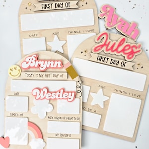 May include: Wooden first day of school boards with names in pink and white. Each board has space for the date, age, and things they love. Additional boards include a rainbow, clouds, and a pencil.