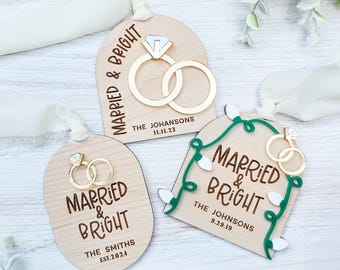 May include: Three wooden ornaments featuring the text "Married & Bright" and a pair of gold rings. The ornaments are decorated with a green garland, white lights, and a diamond ring.