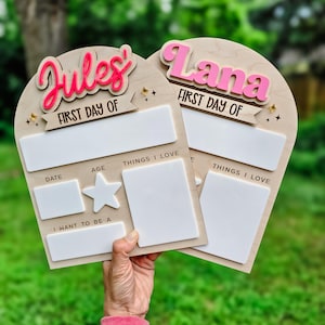 May include: Two wooden first day of school signs with the names "Jules" and "Lana" in pink and red. Each sign has space for the date, age, and things they love. A star and the words "I want to be a" are also included.