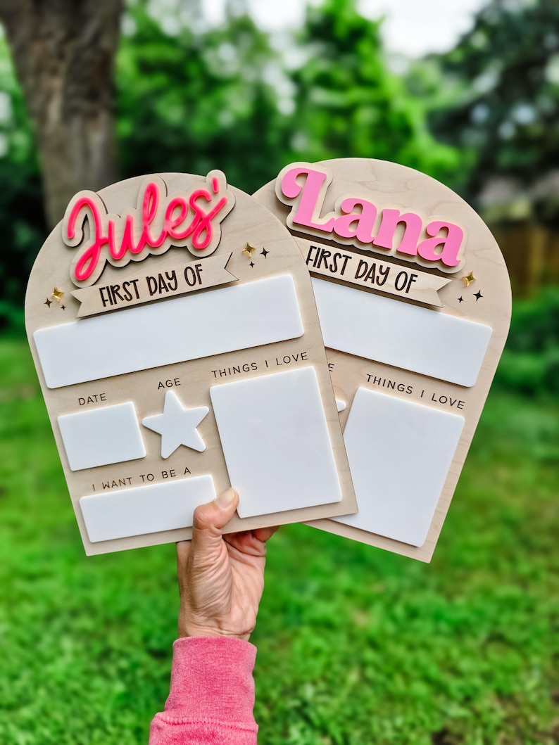 May include: Two wooden first day of school signs with the names "Jules" and "Lana" in pink and red. Each sign has space for the date, age, and things they love. A star and the words "I want to be a" are also included.