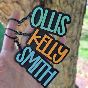 May include: Three rectangular black keychains with rounded corners, each featuring raised lettering in a different color. The top keychain displays the name OLLIS in mint green, the middle displays KELLY in orange, and the bottom displays SMITH in light blue.