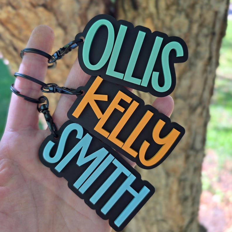May include: Three rectangular black keychains with rounded corners, each featuring raised lettering in a different color. The top keychain displays the name OLLIS in mint green, the middle displays KELLY in orange, and the bottom displays SMITH in light blue.