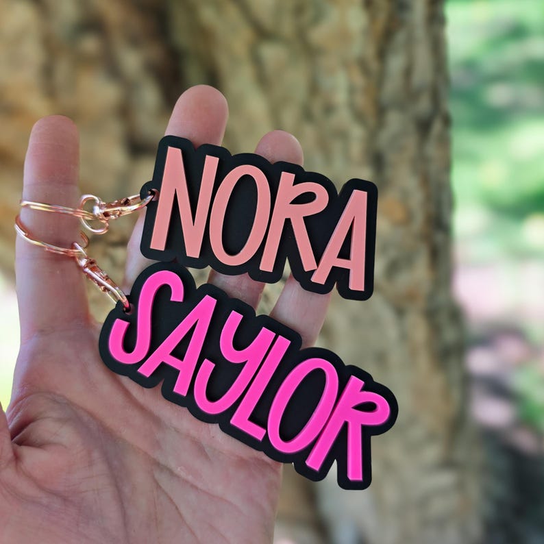 以下が含まれることがあります： NORAとSAYLORの名前が太字の大文字で刻まれた、2つのカスタムアクリルキーホルダー。NORAのキーホルダーは黒い背景にライトピンクの文字、SAYLORのキーホルダーは黒い背景に鮮やかなピンクの文字がデザインされています。