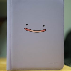 May include: A light purple Ultra Pro binder featuring a cartoon face with black eyes and a smiling mouth. The binder is designed to hold trading cards and has a zipper closure. The Ultra Pro logo is visible on the bottom right corner.