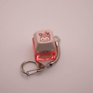 May include: A custom keycap with a cartoon character design, attached to a keychain. The keycap is light pink with a red base, and the keychain is silver. The cartoon character has pink accents.