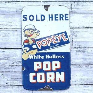 May include: Vintage Popeye Brand popcorn sign with a white background and blue border. The sign features Popeye the Sailor Man and the text "SOLD HERE" and "White Hulless Pop Corn."