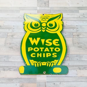 Puede incluir: Letrero vintage de Wise Potato Chips con forma de búho. El letrero es verde con detalles y letras amarillas. El texto "WISE POTATO CHIPS" se muestra de forma destacada. El letrero tiene dos agujeros en la parte superior para colgarlo.