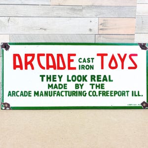 May include: Rectangular vintage sign with a white background and a green border. The sign reads "ARCADE TOYS" in red, with "CAST IRON" in smaller text. Below, it states "THEY LOOK REAL MADE BY THE ARCADE MANUFACTURING CO. FREEPORT ILL."