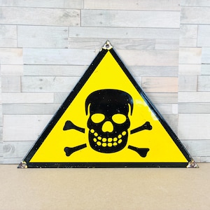 Puede incluir: Un letrero triangular amarillo con un borde negro y un símbolo negro de calavera y huesos cruzados. El letrero es un símbolo de advertencia, que probablemente indica una sustancia o área peligrosa. El letrero es de metal.