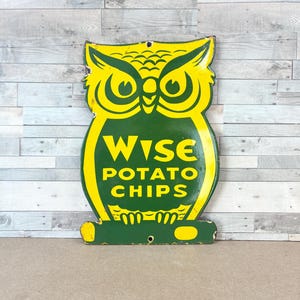 May include: A die-cut metal sign shaped like an owl with a yellow and green color scheme. The sign features the text WISE POTATO CHIPS in yellow lettering on a green background. It includes two small circular holes for mounting at the top and bottom.
