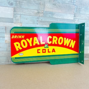 May include: A rectangular metal sign with a green border and a red and yellow center. The text reads DRINK ROYAL CROWN COLA with an RC logo in the middle. The sign features a perpendicular mounting flange on the right side with pre-drilled holes.