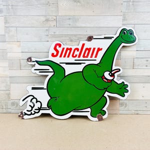 May include: A vintage-style metal sign featuring the green Sinclair dinosaur mascot running. The white background includes the red text Sinclair, black motion lines, and a small red and white cup held by the dinosaur. The sign has visible rust.