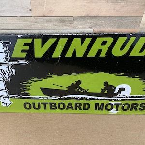 May include: Vintage Evinrude Outboard Motors sign. The sign is black with green lettering and a graphic of a boat with two people. The sign also features a white outline of an outboard motor on the left side.