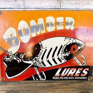 May include: Vintage metal sign with the word "BOMBER" in large silver letters. The sign features a detailed illustration of a fishing lure with the word "LURES" and the tagline "Make the big ones surrender."