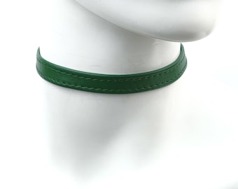 Könnte beinhalten: Ein grünes Leder-Choker-Halsband. Der Choker hat eine kräftige, grüne Farbe und scheint aus glattem Leder zu bestehen. Die Halskette wird auf einem weißen Schaufensterkopf präsentiert.