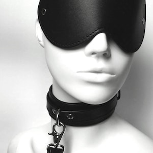 Può includere: Benda nera in pelle con bordo cucito e un anello in metallo argentato su ogni lato. Un collare in pelle nera con dettagli in metallo argentato è attaccato a un guinzaglio in pelle nera con un moschettone in metallo argentato.