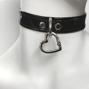 Puede incluir: Un collar de cuero negro con un dije de corazón plateado. El collar tiene un acabado brillante y está adornado con tachuelas plateadas.