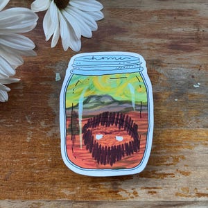 May include: A sticker featuring a jar with a landscape painting inside. The jar has a blue lid with the word "home" written on it. The painting inside depicts a yellow and green sky, mountains, and a brown object.