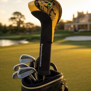 Puede incluir: Una funda para palo de golf con forma de cobra, textura de piel de serpiente amarilla y negra, ojos verdes y un cuello negro con el texto blanco BigstickDiplomacy, colocada en una bolsa de golf negra con varios palos de metal visibles.