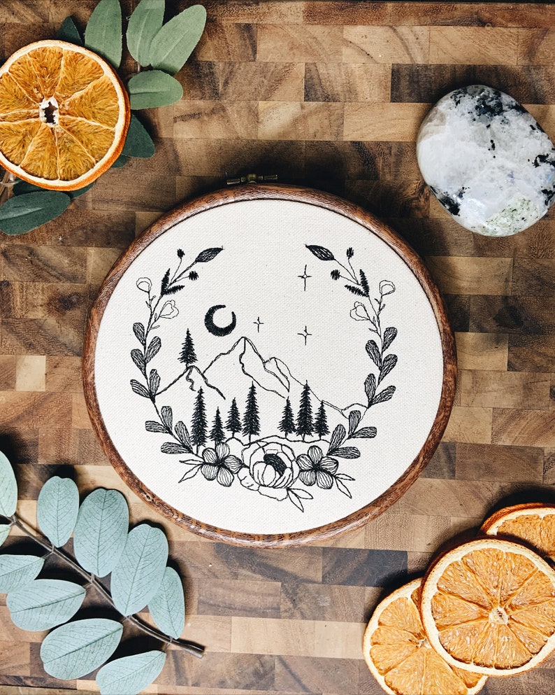 May include: A circular wooden embroidery hoop containing white fabric with a black thread design of a mountain range, pine trees, a crescent moon, and stars, all framed by a floral and leaf wreath. The embroidery is centered on the fabric.