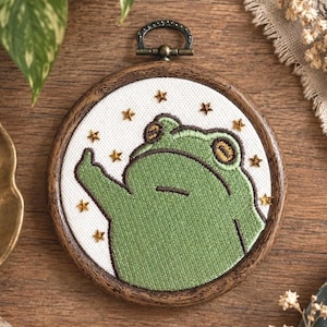 May include: A circular embroidery hoop featuring a green frog with a flat expression, one arm raised, and small gold star accents on a white fabric background. The hoop is made of dark wood and includes a metal hanging loop at the top.