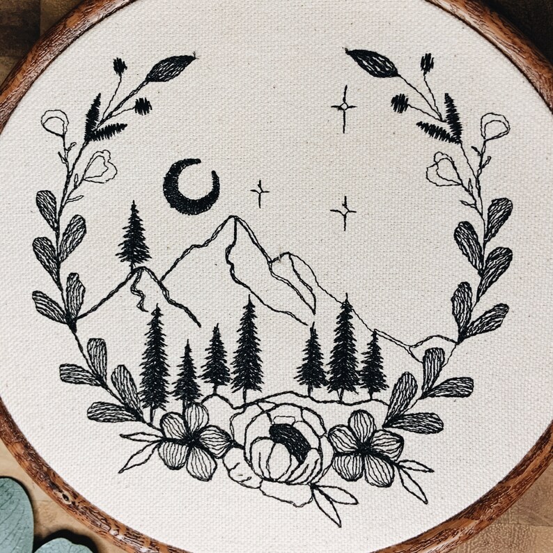 May include: A circular embroidery hoop containing off-white fabric with a black thread design. The embroidery features a mountain range, pine trees, a crescent moon, and stars, all framed by a wreath of leaves and flowers at the base.