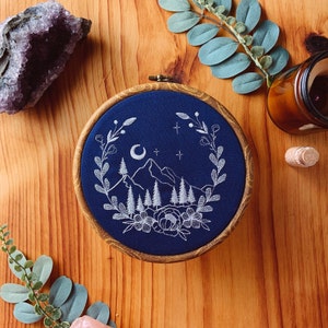 May include: A circular wooden embroidery hoop containing dark blue fabric with white thread stitching. The design features a mountain range, pine trees, a crescent moon, stars, and a floral wreath border surrounding the central landscape scene.