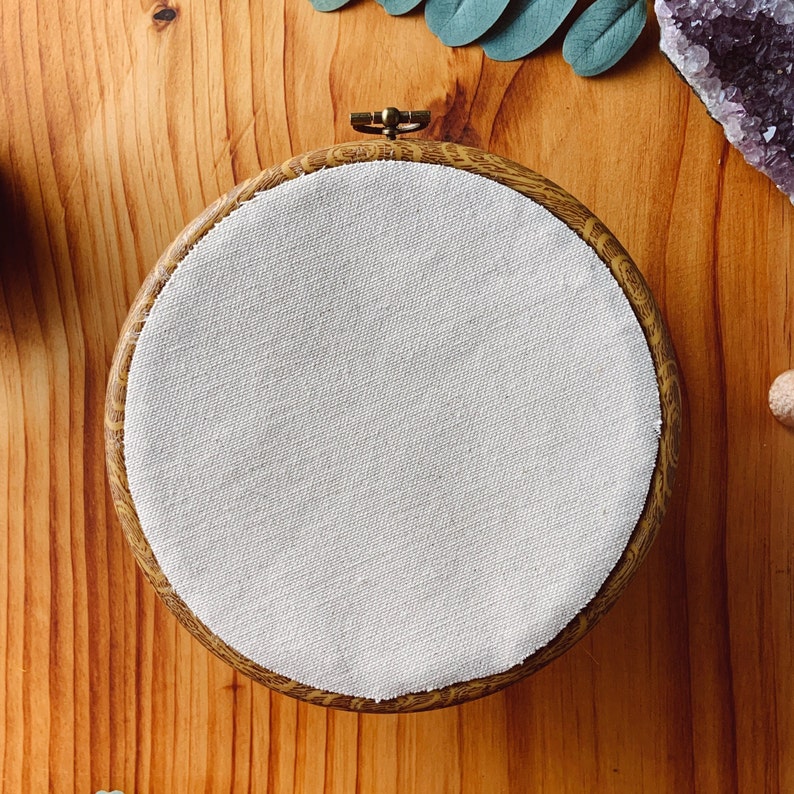 May include: A circular embroidery hoop featuring a faux wood grain pattern on the outer frame and a metallic tension screw at the top. The hoop is tightly fitted with a plain, off-white, unbleached fabric stretched across the entire circular opening.