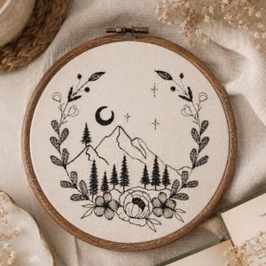 May include: A circular wooden embroidery hoop containing off-white fabric with a black thread design. The embroidery features a mountain range, pine trees, a crescent moon, and stars, all framed by a wreath of leaves and flowers.