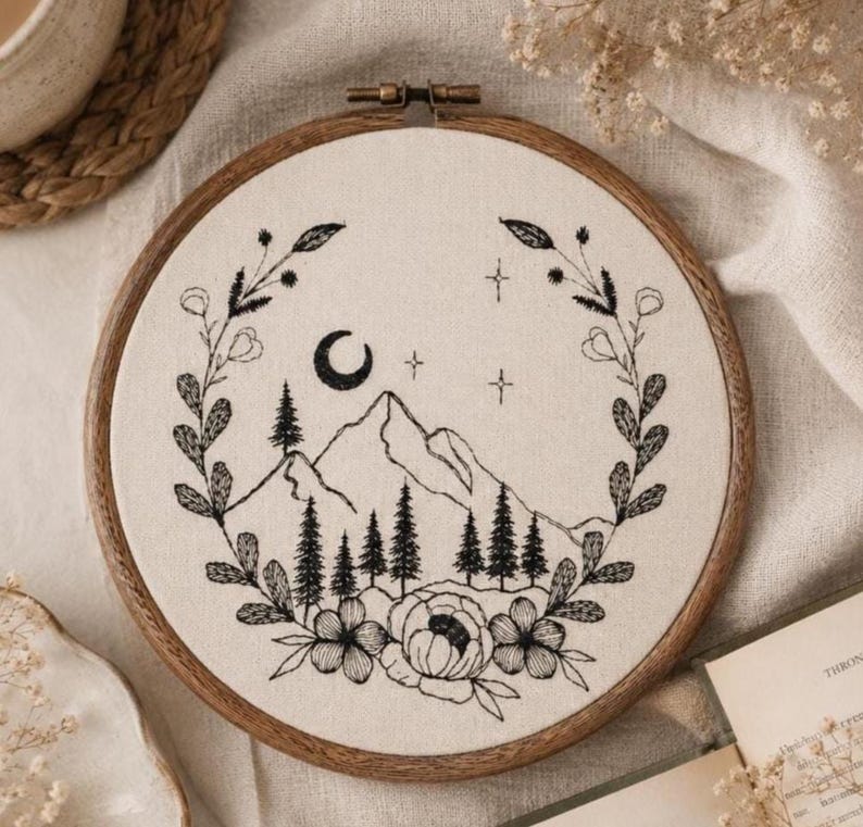 May include: A circular wooden embroidery hoop containing off-white fabric with a black thread design. The embroidery features a mountain range, pine trees, a crescent moon, and stars, all framed by a wreath of leaves and flowers.