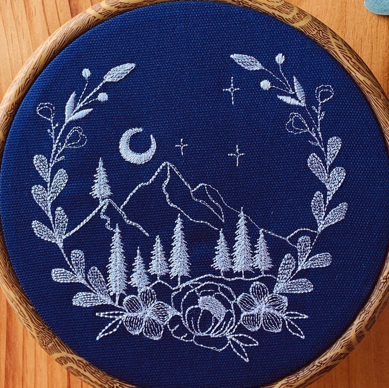 May include: A circular embroidery hoop containing dark blue fabric with white thread stitching. The design features a mountain range, pine trees, a crescent moon, and stars, all framed by a wreath of leaves and flowers at the bottom.