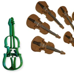 May include: A green plastic violin-shaped cookie cutter next to several brown violin-shaped cookies. The cookies have black and white details, resembling the strings and bridge of a violin. The image is on a white background.