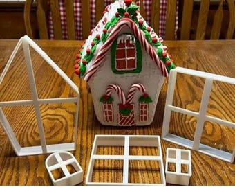 Puede incluir: Una casa de jengibre decorada con detalles de bastones de caramelo y un techo rojo y verde, rodeada de moldes de marcos de ventanas de plástico blanco. Tema festivo.