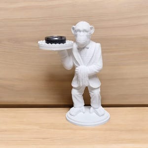 May include: A white monkey figurine in a tuxedo holding a small, round, black object on a decorative tray. The monkey is standing on a round base. The background is a light brown wooden surface.