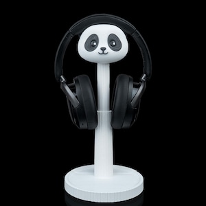 May include: A white panda-shaped headphone stand with black headphones. The stand has a round base and a vertical support topped with a panda head. The headphones are black and rest on the panda's ears.