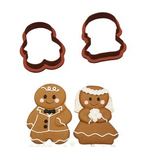 May include: A set of gingerbread man and woman cookie cutters and decorated cookies. The cutters are dark brown, and the cookies are decorated with white icing and pink cheeks. The gingerbread man wears a tuxedo, and the woman wears a veil.