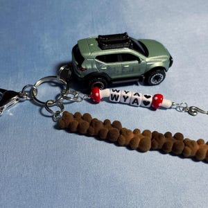 May include: A silver keychain with a brown fuzzy charm, a small green toy car, and white beads with the letters "WYA" and a heart charm.