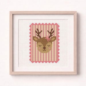 May include: A framed cross-stitch art piece featuring a deer's face with closed eyes and a pink flower. The deer is brown with dark brown antlers, set against a pink and white striped background with a scalloped edge.