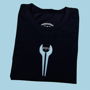 Puede incluir: Una camiseta negra de manga corta con un gráfico bordado de una espada de energía azul claro en el pecho. La etiqueta interior del cuello muestra el texto MOREJAMESON. La prenda está hecha de tela negra sólida con cuello redondo.