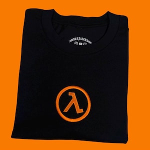 Puede incluir: Una camiseta negra de cuello redondo con un símbolo lambda naranja bordado dentro de un círculo en el pecho. La etiqueta interior del cuello muestra el texto MOREJAMESON junto con pequeños iconos de redes sociales para Instagram, YouTube y Twitch.