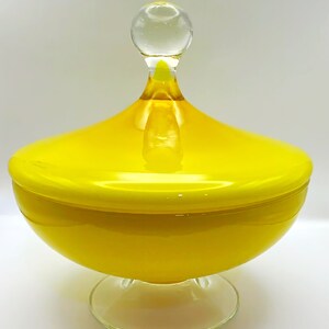 May include: A bright yellow glass candy dish with a matching lid and a clear glass pedestal base. The lid features a smooth, rounded yellow dome topped with a small, clear glass spherical finial, creating a vibrant and polished appearance.