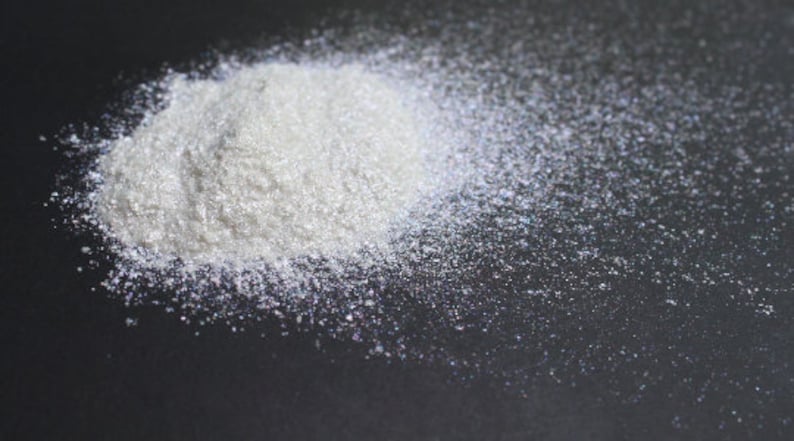 Puede incluir: Un mont&oacute;n de purpurina blanca iridiscente sobre una superficie negra. La purpurina est&aacute; dispersa alrededor del mont&oacute;n, creando un efecto brillante.